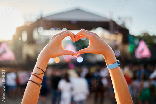 Hands shape heart sign at sunset beach music fest, crowd enjoys live concert. Outdoor summer event, happy fans party, love symbol, festive vibe by the sea. Silhouette, sun glow, entertainment. photo
