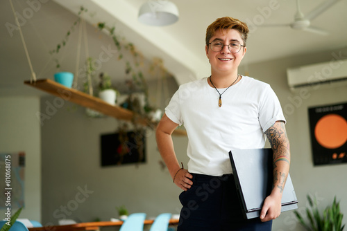 Confident transgender male employee stands with laptop in modern office setup. Smart casual work attire with glasses, tattoos. Pro holds tech, represents workplace inclusion, gender diversity.