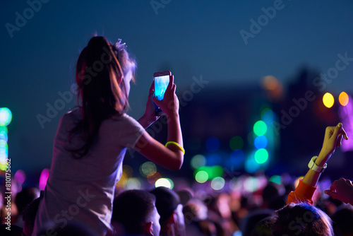 Girl films music concert on smartphone amid lively festival crowd at twilight. Audience enjoys live performance, memories, vibrant stage lights beam. Fans wristbands share experience social media. photo