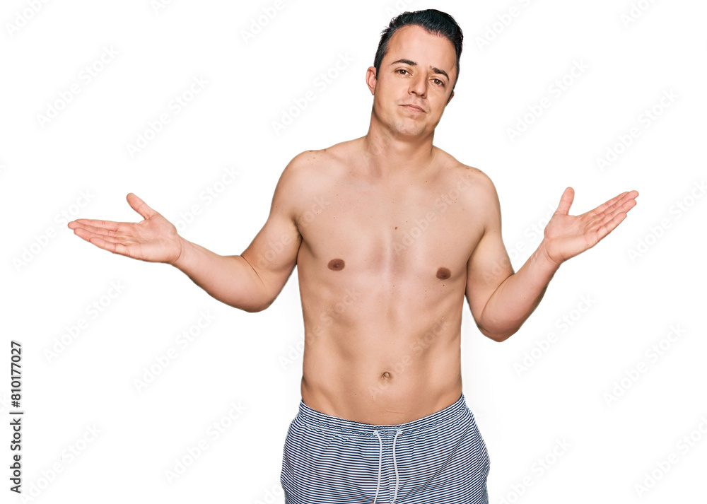 Handsome young man wearing swimwear shirtless clueless and confused expression with arms and hands raised. doubt concept. Handsome young man wearing swimwear shirtless clueless and confused expression with arms and hands raised. doubt concept.