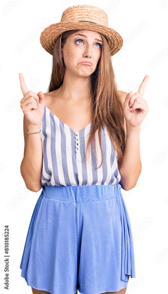 Young beautiful blonde woman wearing summer hat pointing up looking sad and upset, indicating direction with fingers, unhappy and depressed.