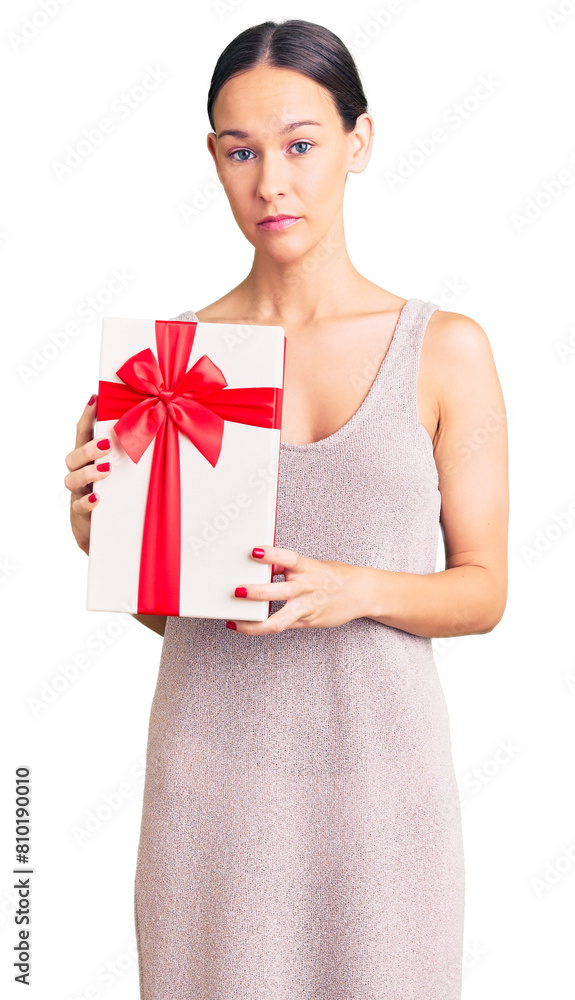 Beautiful brunette young woman holding gift thinking attitude and sober expression looking self confident Beautiful brunette young woman holding gift thinking attitude and sober expression looking self confident