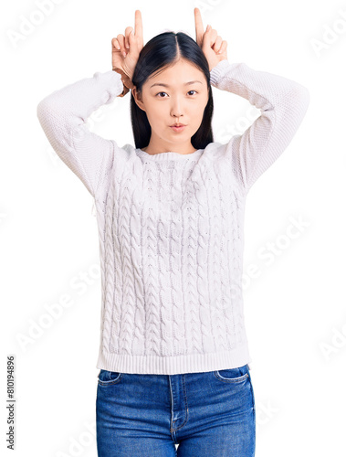 Young beautiful chinese woman wearing casual sweater doing funny gesture with finger over head as bull horns