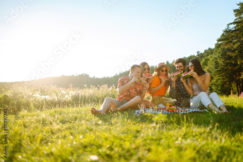 Cheerful friends eating fruits while sitting on the park. Young people having fun in summer vacation. Foods, travel, nature and picnic concept