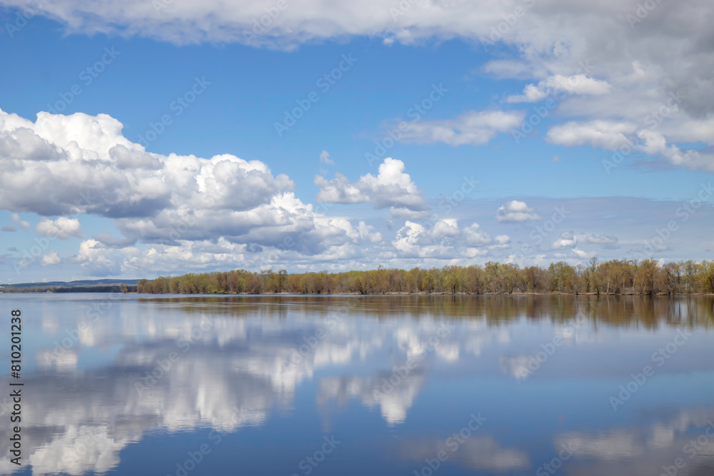 Calm waters with clouds and sky reflected in water, heavy clouds, forested shore in background Calm waters with clouds and sky reflected in water, heavy clouds, forested shore in background