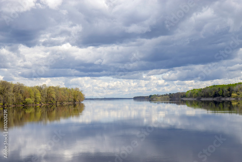 Calm waters with clouds and sky reflected in water, heavy clouds, forested shore in background
