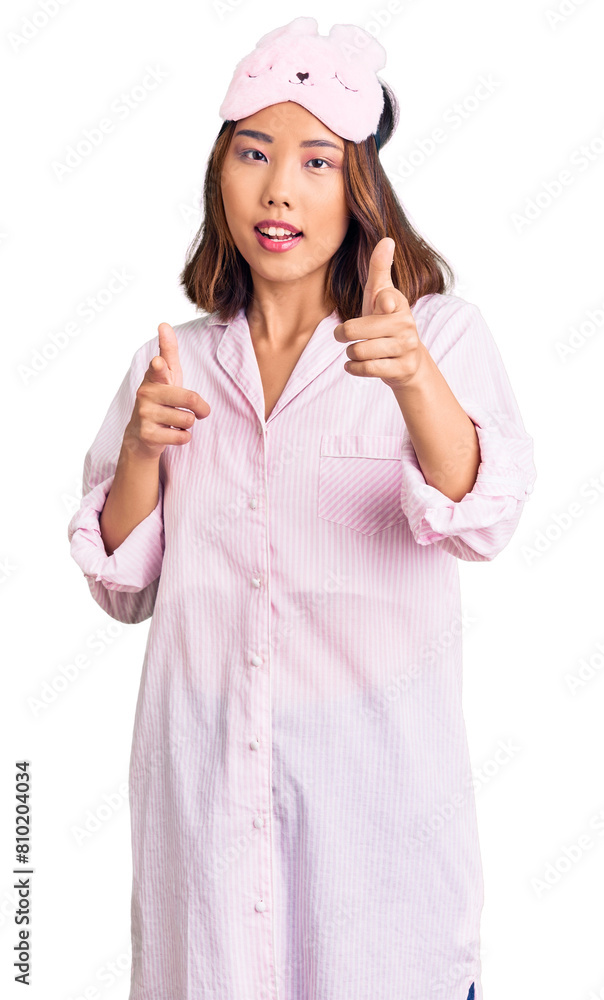 Young beautiful chinese girl wearing sleep mask and pajama pointing fingers to camera with happy and funny face. good energy and vibes. Young beautiful chinese girl wearing sleep mask and pajama pointing fingers to camera with happy and funny face. good energy and vibes.