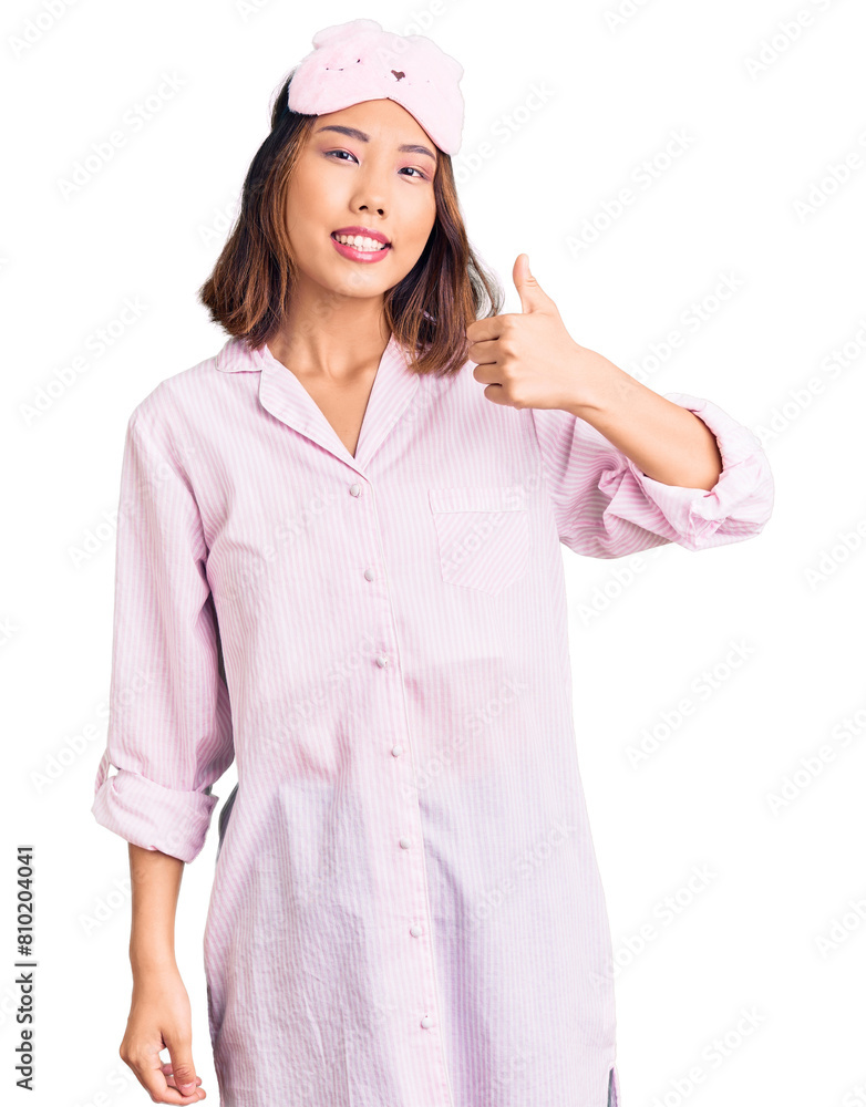 Young beautiful chinese girl wearing sleep mask and pajama doing happy thumbs up gesture with hand. approving expression looking at the camera showing success. Young beautiful chinese girl wearing sleep mask and pajama doing happy thumbs up gesture with hand. approving expression looking at the camera showing success.