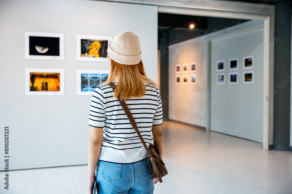 © sorapop - Asian young woman standing she looking art gallery in front of colorful framed paintings pictures on white wall, female watch at photo frame to leaning against at exhibit museum, Back view