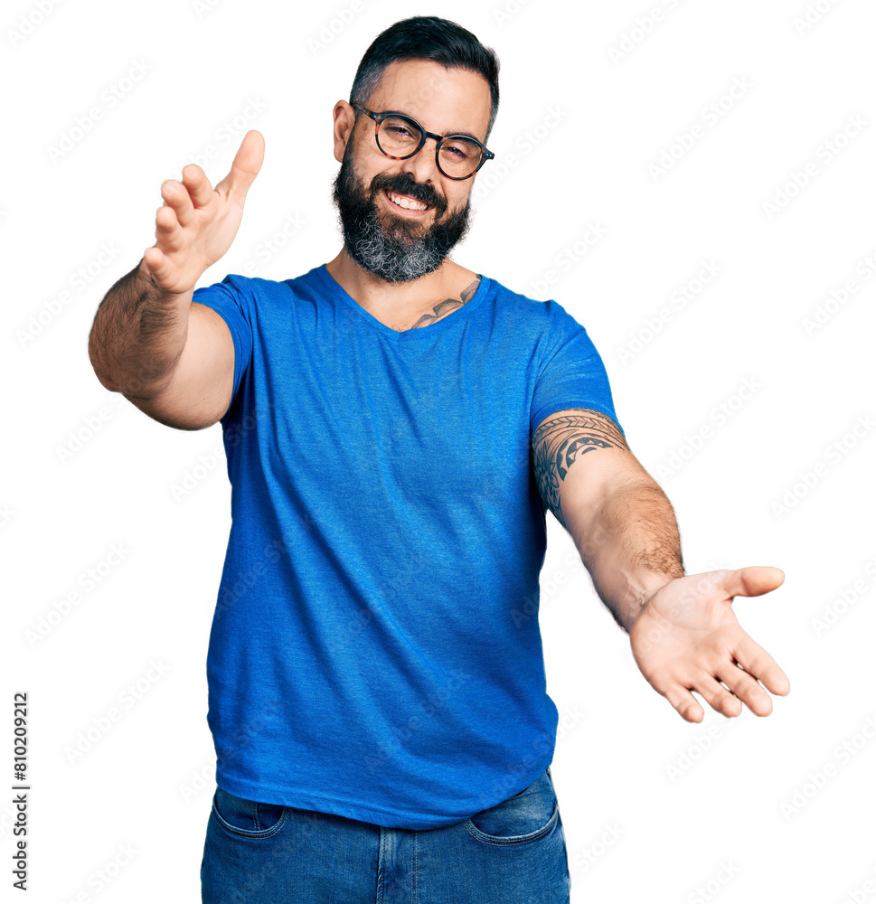 Hispanic man with beard wearing casual t shirt and glasses looking at the camera smiling with open arms for hug. cheerful expression embracing happiness. Hispanic man with beard wearing casual t shirt and glasses looking at the camera smiling with open arms for hug. cheerful expression embracing happiness.