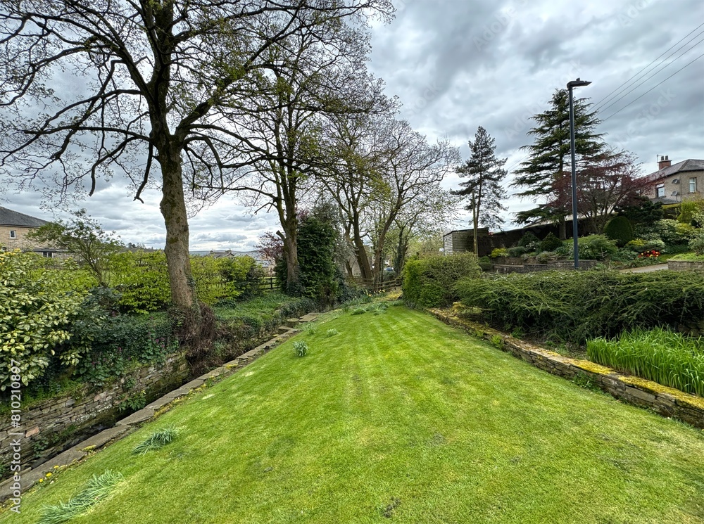 Obraz premium A quaint green area with a gently sloping lawn, an array of plants and bushes, and a stream bordered by old trees, in the charming village of Bradley, Yorkshire, UK.