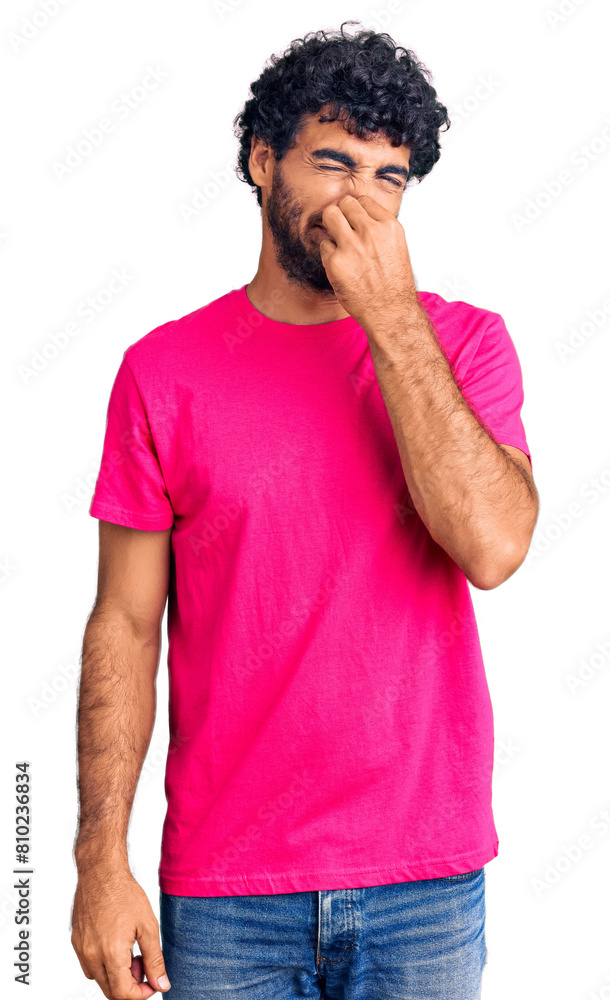 Handsome young man with curly hair and bear wearing casual pink tshirt smelling something stinky and disgusting, intolerable smell, holding breath with fingers on nose. bad smell Handsome young man with curly hair and bear wearing casual pink tshirt smelling something stinky and disgusting, intolerable smell, holding breath with fingers on nose. bad smell