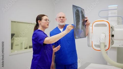 A confident brunette girl with glasses in a blue uniform communicates together and consults with a confident man doctor with gray hair while summing up the results of fluorography and confirming the photo
