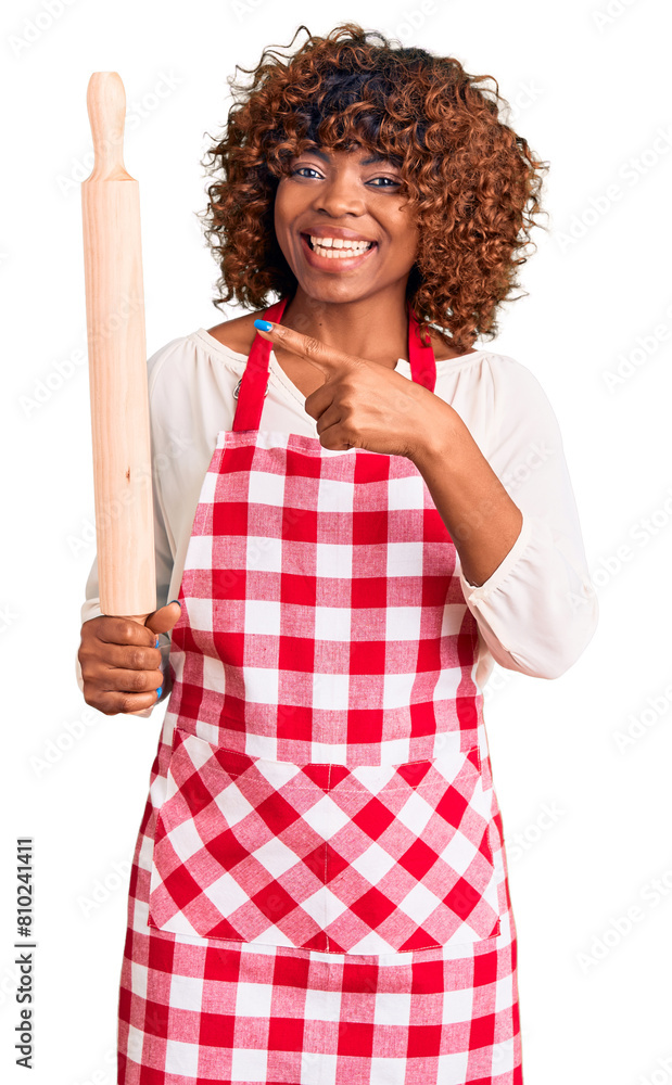 Young african american woman wearing apron holding rolling pin smiling happy pointing with hand and finger Young african american woman wearing apron holding rolling pin smiling happy pointing with hand and finger