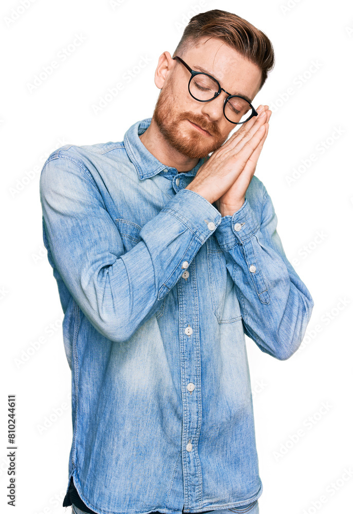 Young redhead man wearing casual denim shirt sleeping tired dreaming and posing with hands together while smiling with closed eyes. Young redhead man wearing casual denim shirt sleeping tired dreaming and posing with hands together while smiling with closed eyes.