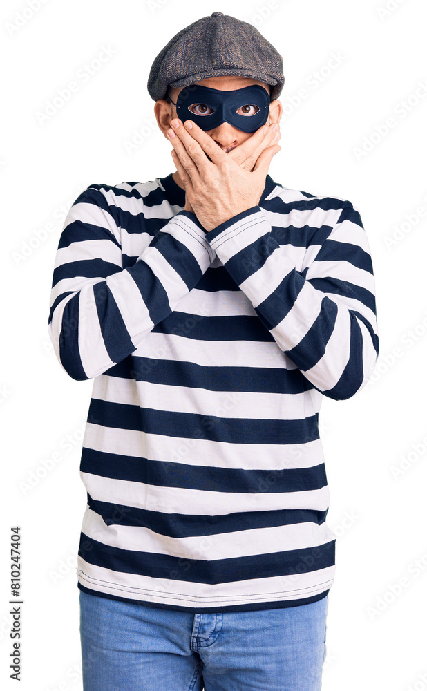 Young handsome man wearing burglar mask shocked covering mouth with hands for mistake. secret concept. Young handsome man wearing burglar mask shocked covering mouth with hands for mistake. secret concept.