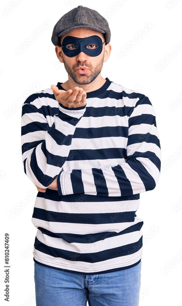 Young handsome man wearing burglar mask looking at the camera blowing a kiss with hand on air being lovely and sexy. love expression. Young handsome man wearing burglar mask looking at the camera blowing a kiss with hand on air being lovely and sexy. love expression.