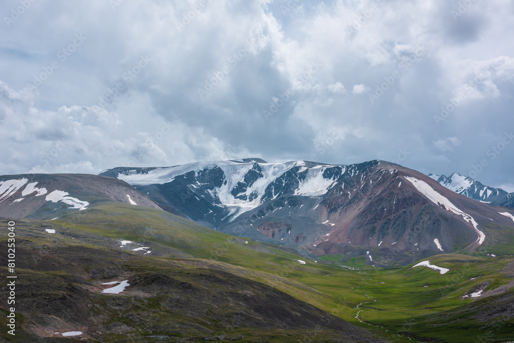 Dramatic view to large snow-capped mountain range under gray rainy cloudy sky. Shadows of clouds on green valley and stony hills. Snowy rocky mountain wall. Changeable weather in big snow mountains.