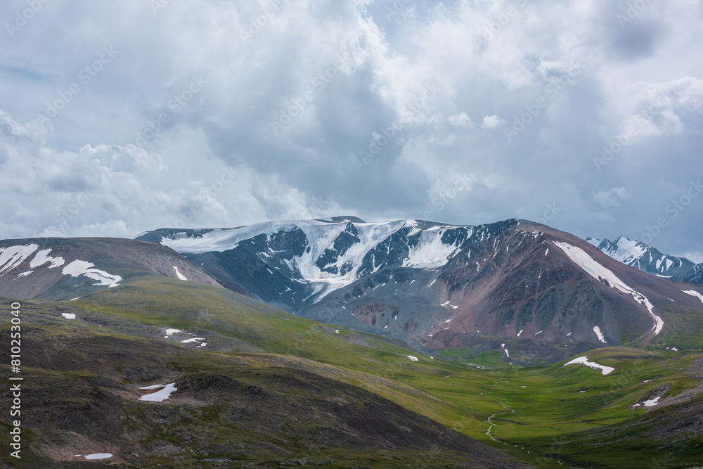 Dramatic view to large snow-capped mountain range under gray rainy cloudy sky. Shadows of clouds on green valley and stony hills. Snowy rocky mountain wall. Changeable weather in big snow mountains. Dramatic view to large snow-capped mountain range under gray rainy cloudy sky. Shadows of clouds on green valley and stony hills. Snowy rocky mountain wall. Changeable weather in big snow mountains.