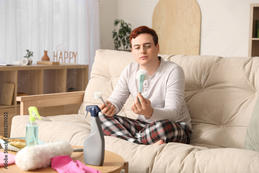 Fototapeta premium Young man holding cleaning brushes on sofa, home chores and domestic responsibilities concept, housekeeping tools