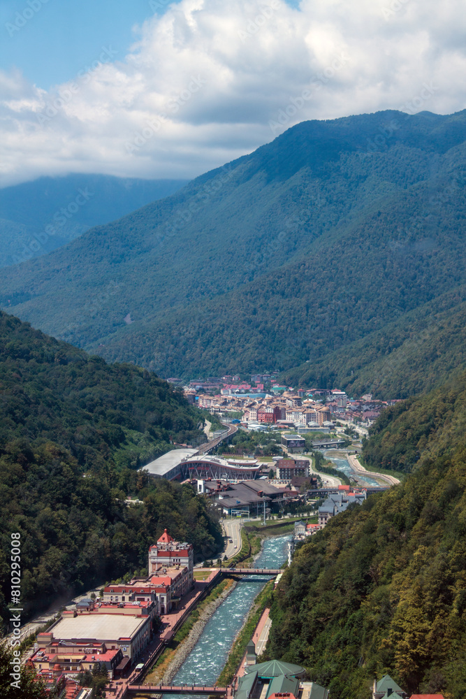 Fototapeta premium Panoramic top view of Krasnaya Polyana among high green mountains and white clouds on a sunny summer day