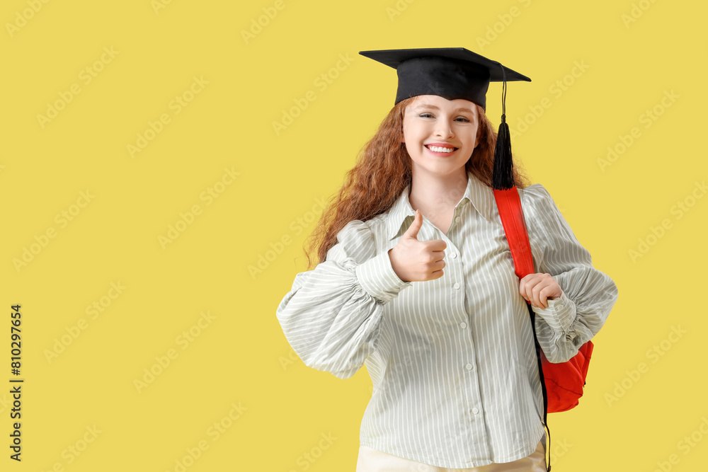 Happy female student in graduation hat with backpack showing thumb-up on yellow background Happy female student in graduation hat with backpack showing thumb-up on yellow background