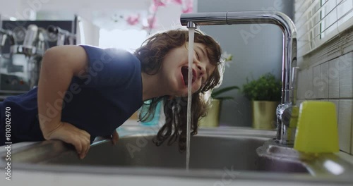 Cute boy with curly hair leans over a kitchen sink to drink water straight from the tap, with daylight illuminating the scene.