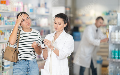 Young female customer complaining of ache speaking to woman apothecary holding box of medicine in drugstore