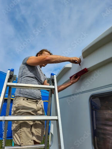 An experienced handyman is meticulously repairing a roof vent on a recreational vehicle using a screwdriver. He is wearing casual work attire and the clear blue sky is visible in the background.