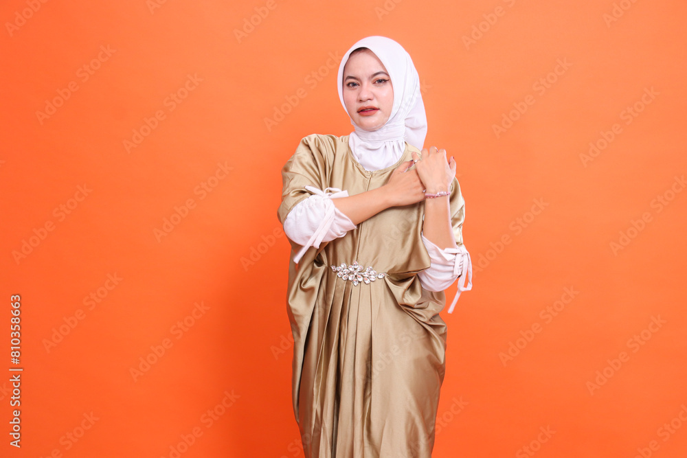 gesture of a beautiful mature Asian woman, sad, both hands holding her sore shoulders, wearing a kaftan Muslim dress with a hijab, isolated on an orange background. Concept of health, education gesture of a beautiful mature Asian woman, sad, both hands holding her sore shoulders, wearing a kaftan Muslim dress with a hijab, isolated on an orange background. Concept of health, education
