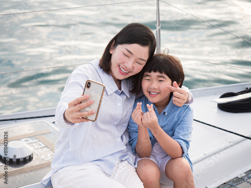 Asian mother and son taking a selfie with smartphone on sailing yacht together on summer vacation, leisure recreation fun travel family lifestyle.