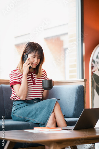 Young woman balances a phone conversation and coffee while working on her laptop in a relaxed urban cafe setting.