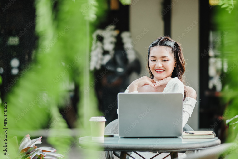 Cheerful young freelancer engaged with her laptop at a peaceful outdoor cafe surrounded by plants. Cheerful young freelancer engaged with her laptop at a peaceful outdoor cafe surrounded by plants.
