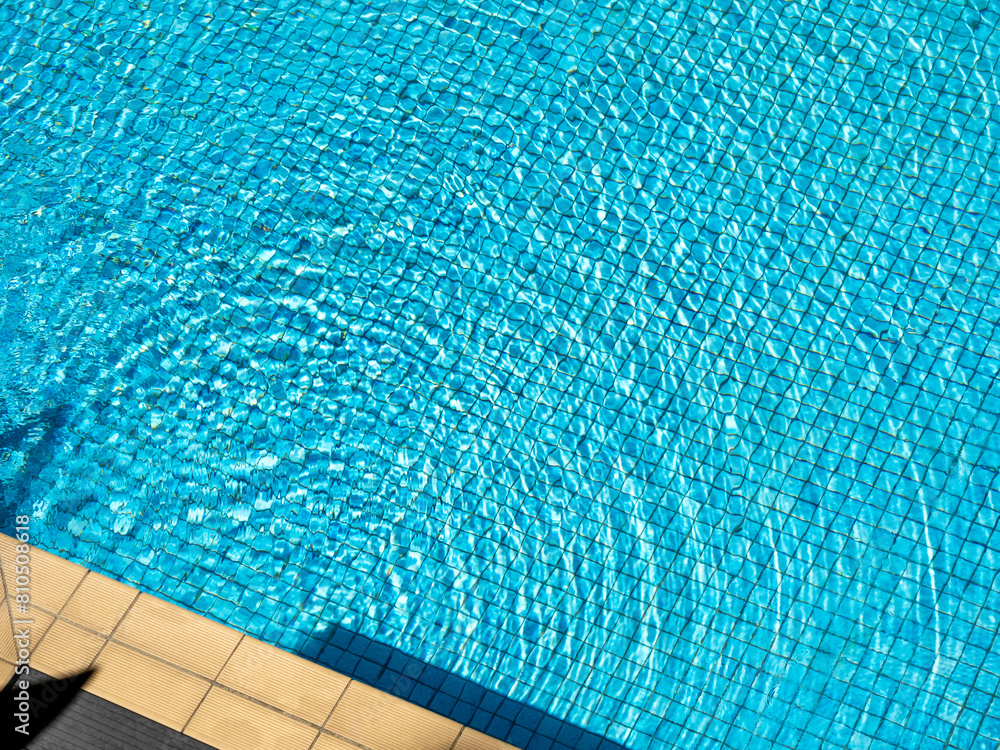 Top view space of blurred round rippled clear water moving on blue grid mosaic tiles in swimming pool background, near pool edge. Empty space on poolside on sunny day, summer background.