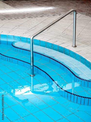 Chrome metal grab bar on swimming pool, curved steps with clear water surface with no body, vertical style. Abstract pool texture, underwater pattern blue grid tiles, no people. Summer background.