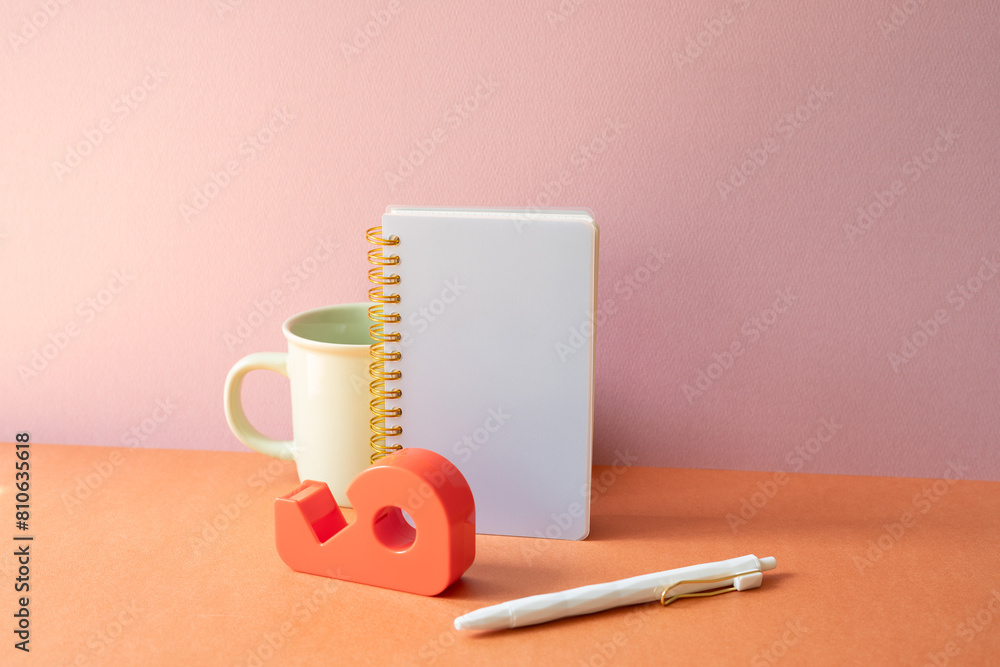 Workspace office desk with notebook, mug cup, pen, tape dispenser. pink orange background