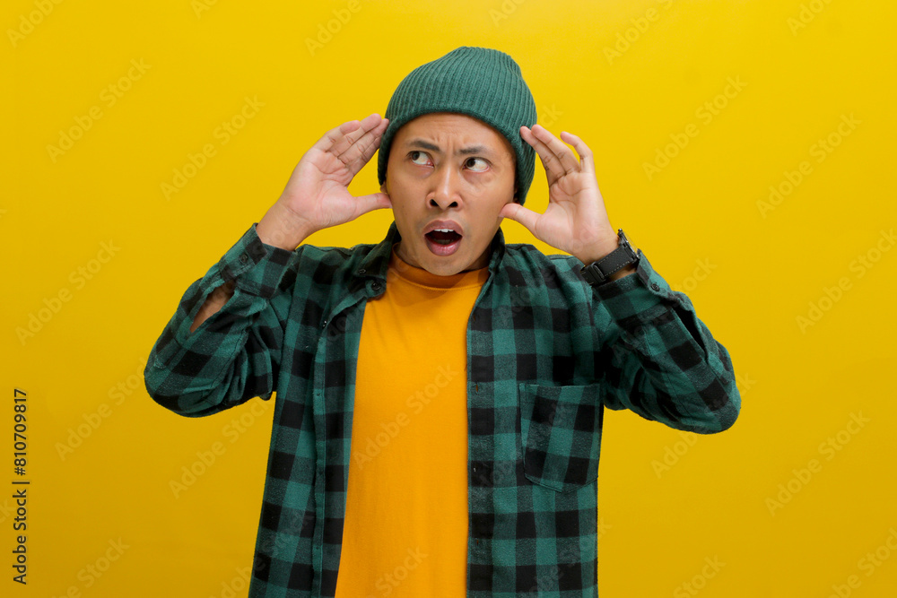 Shocked and anxious young Asian man, dressed in a beanie hat and casual shirt, appears panicked, holding his hands on his head while standing against a yellow background Shocked and anxious young Asian man, dressed in a beanie hat and casual shirt, appears panicked, holding his hands on his head while standing against a yellow background