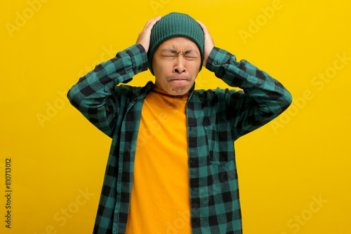 Stressed young Asian man dressed in a beanie hat and casual shirt is holding his head, possibly indicating feelings of overwhelm or distress , isolated on yellow background. © Jamaludinyusup