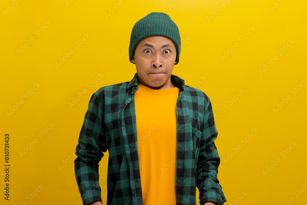 A worried young Asian student, dressed in a beanie hat and casual shirt, appears anxious and fearful about facing a challenging question during the final exam. Standing against yellow background. A worried young Asian student, dressed in a beanie hat and casual shirt, appears anxious and fearful about facing a challenging question during the final exam. Standing against yellow background.