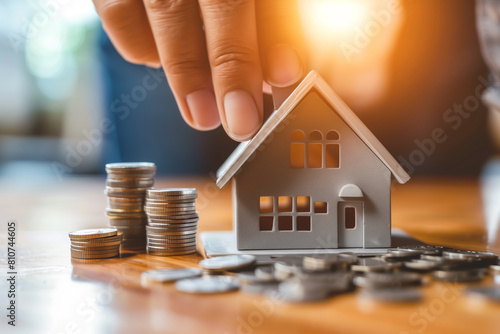 Hand placing a miniature house among stacked coins on a wooden surface, Property investment, real estate savings, and home ownership concept.