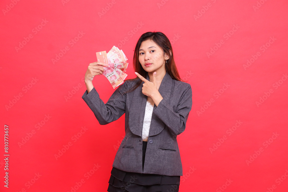 The expression of an elegant Asian office girl holding a credit, debit card and money while pointing it at the camera wearing a gray jacket and skirt on a red background. for transaction advertising The expression of an elegant Asian office girl holding a credit, debit card and money while pointing it at the camera wearing a gray jacket and skirt on a red background. for transaction advertising
