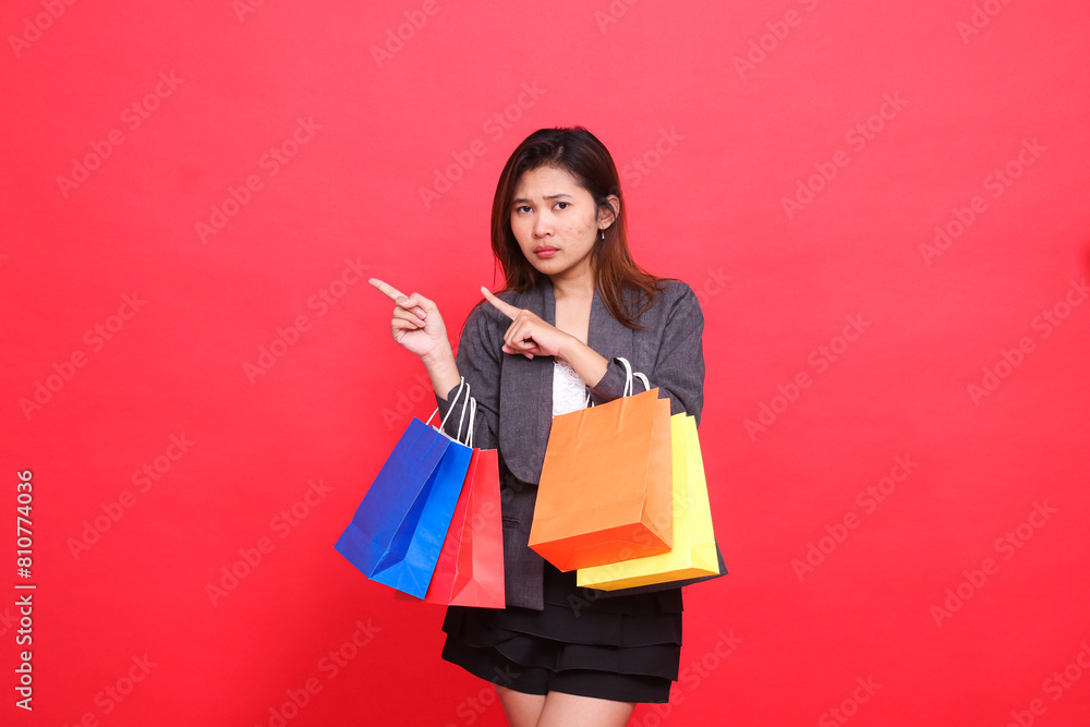 Fototapeta premium the expression of an annoyed indonesia office woman carrying a shopping paper bag with her right hand pointing up wearing a formal jacket on a red background. for commercial, lifestyle and sales