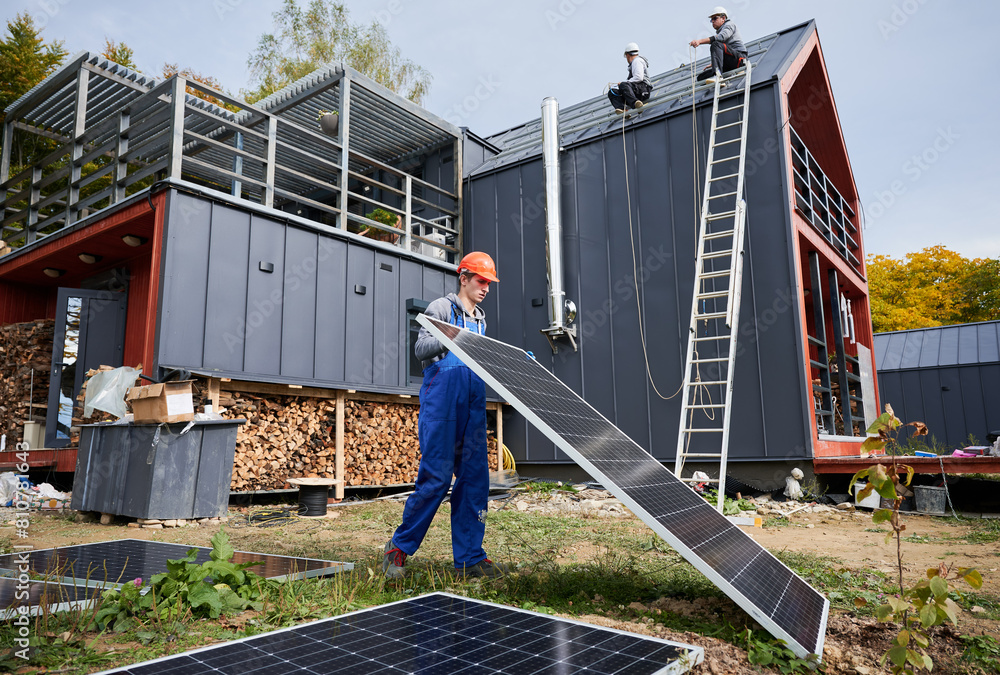 Roofers installing solar panel system on roof of house. Man worker in helmet carrying photovoltaic solar module outdoors. Concept of alternative and renewable energy. Roofers installing solar panel system on roof of house. Man worker in helmet carrying photovoltaic solar module outdoors. Concept of alternative and renewable energy.