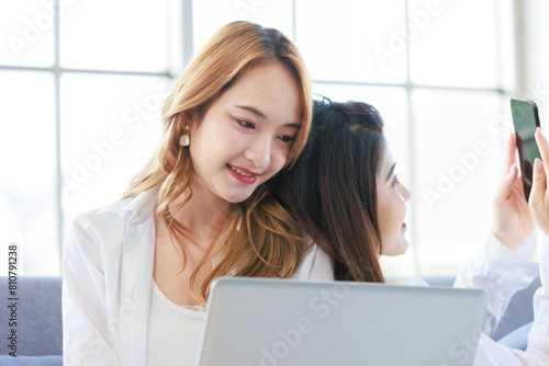 Two asian happy young woman casual outfit sitting on cozy sofa by the window in living room, she smiling looking smartphone and laptop computer at home, spending weekend resting relaxing and enjoying