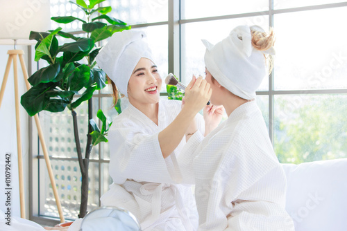 Joyful two asian happy beautiful young women smiling wearing bathrobes and wrapped in towels applying makeup, making and enjoy having fun on bed in bedroom at home