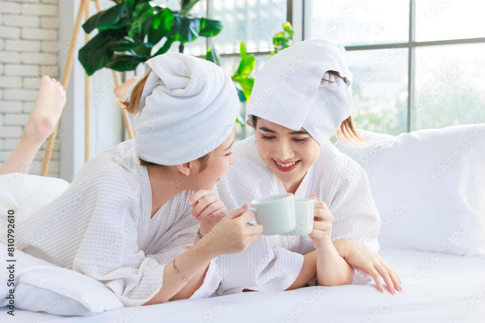 Asian happy beautiful young woman smiling wearing a towel and a white bathrobes, drinking coffee and having a conversation on bed, Two female friends spending time together at home, spa concept. Asian happy beautiful young woman smiling wearing a towel and a white bathrobes, drinking coffee and having a conversation on bed, Two female friends spending time together at home, spa concept.