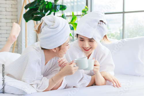Asian happy beautiful young woman smiling wearing a towel and a white bathrobes, drinking coffee and having a conversation on bed, Two female friends spending time together at home, spa concept.