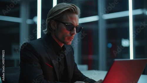 Businessman working on laptop in the office, Handsome young man with blond back brushed hair wearing black suit and sunglasses looking like the main antagonist or protagonist of a thriller movie  photo