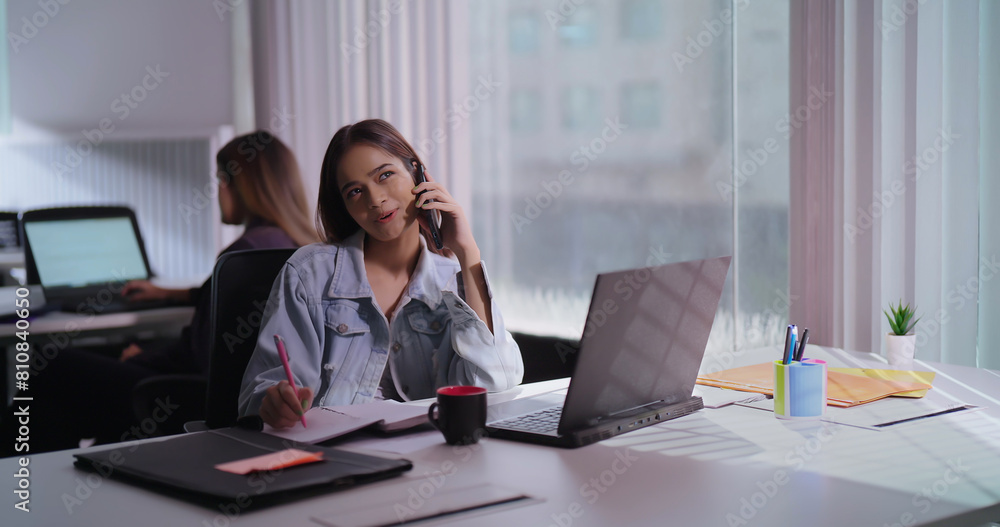 Obraz premium Asian India teen age staff girl sitting desk chair use laptop hold mobile phone talk client help online indoor office Indian busy young adult gen z woman lady use device call centre do job work task
