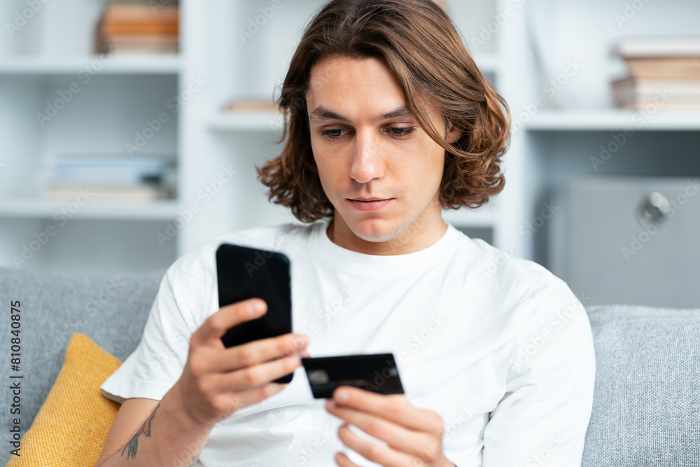 Young Man Shopping Online, Caucasian Male Using Credit Card And Smartphone On Couch, Focused Expression, Indoor Setting, Personal Finance, E-Commerce, Technology Young Man Shopping Online, Caucasian Male Using Credit Card And Smartphone On Couch, Focused Expression, Indoor Setting, Personal Finance, E-Commerce, Technology